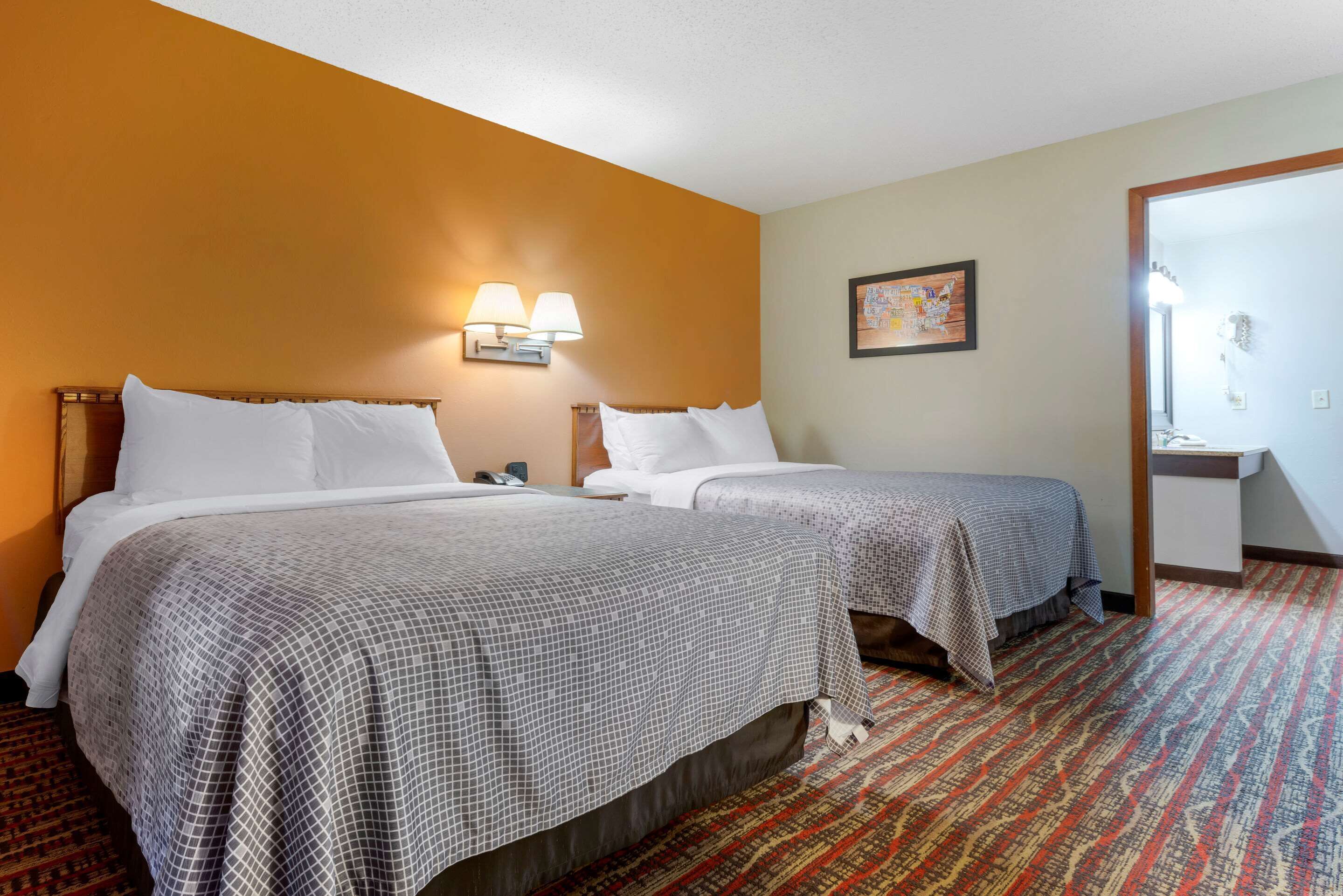 A hotel room with two beds, a patterned carpet, a wall-mounted lamp, and a framed picture.