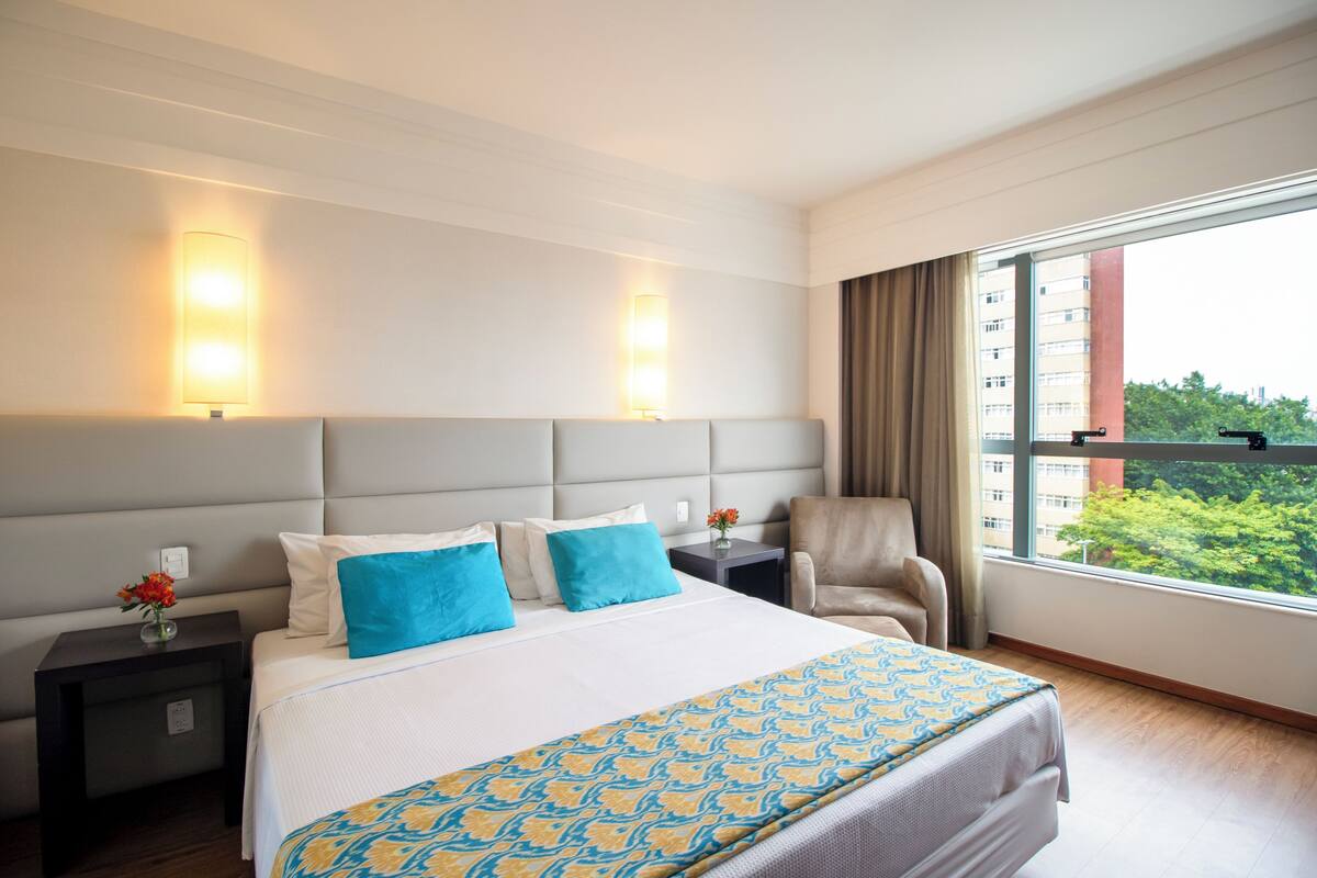 Habitación de hotel con una cama grande, dos lámparas de noche, una silla, una mesita de noche con un jarrón de flores y una ventana con vistas a edificios y árboles.