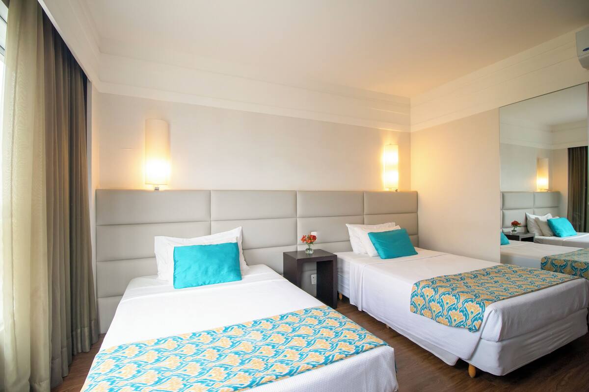 Una habitación de hotel con dos camas, cada una con una colcha estampada, una mesita de noche con un jarrón de flores y un espejo grande en la pared.