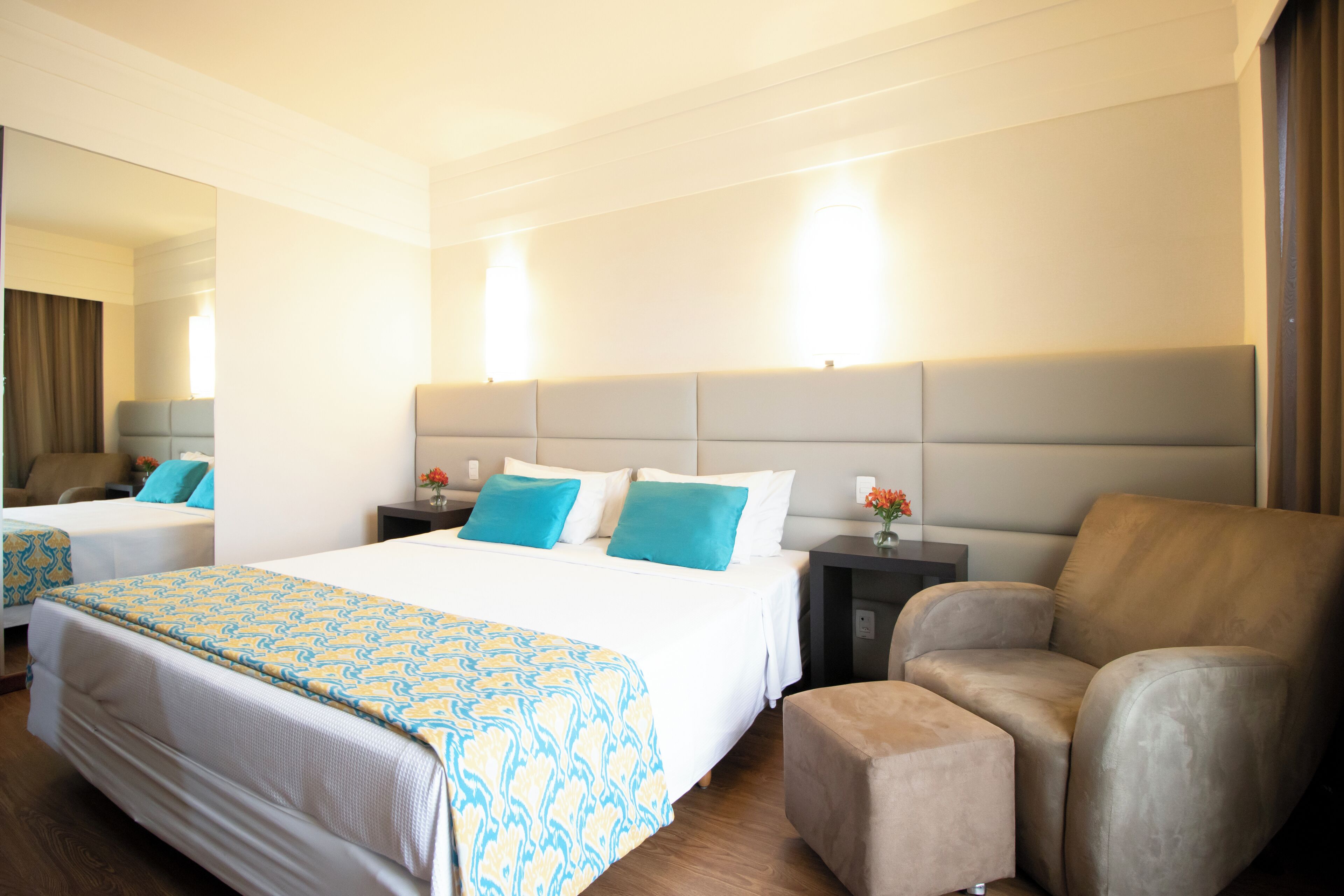 Quarto de hotel com duas camas, um espelho grande, um sofá e uma mesinha de cabeceira com um vaso de flores.