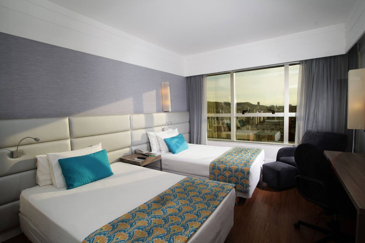 Habitación de hotel con dos camas, un escritorio y un ventanal con vistas a la ciudad.