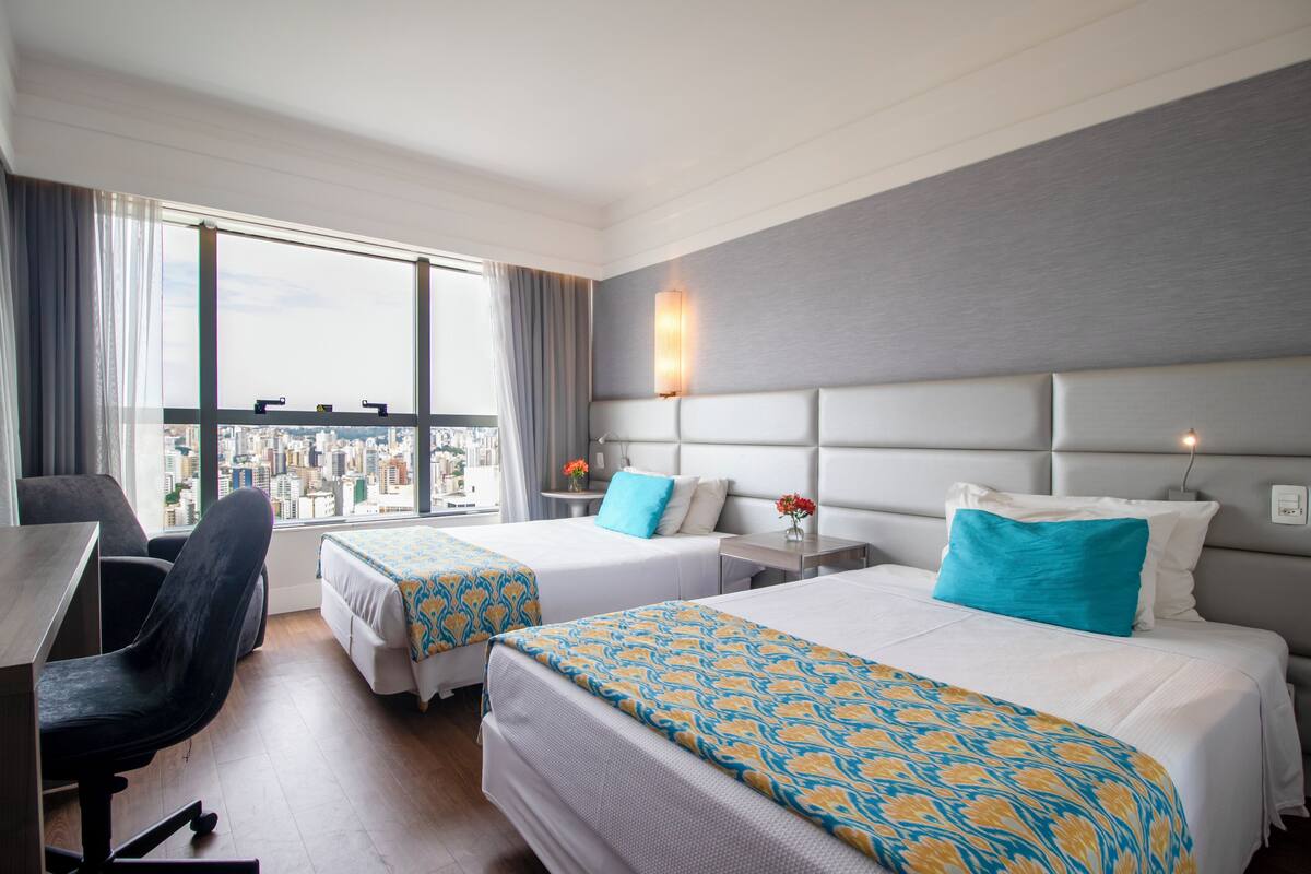 Habitación de hotel con dos camas, un escritorio y una silla. Amplia ventana con vista a la ciudad.