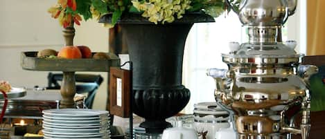A dining table with a floral tablecloth, a silver samovar, and a vase with flowers.