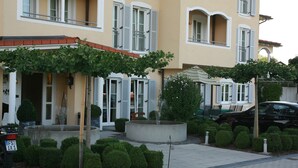 Un bâtiment à la façade jaune, aux volets blancs et à l’entrée pavée, agrémentée d’une fontaine et de buissons taillés.