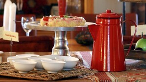 Une table à manger avec une théière rouge, des bols blancs et un gâteau présenté sur un présentoir. On aperçoit des bouteilles sur des étagères à l’arrière-plan.