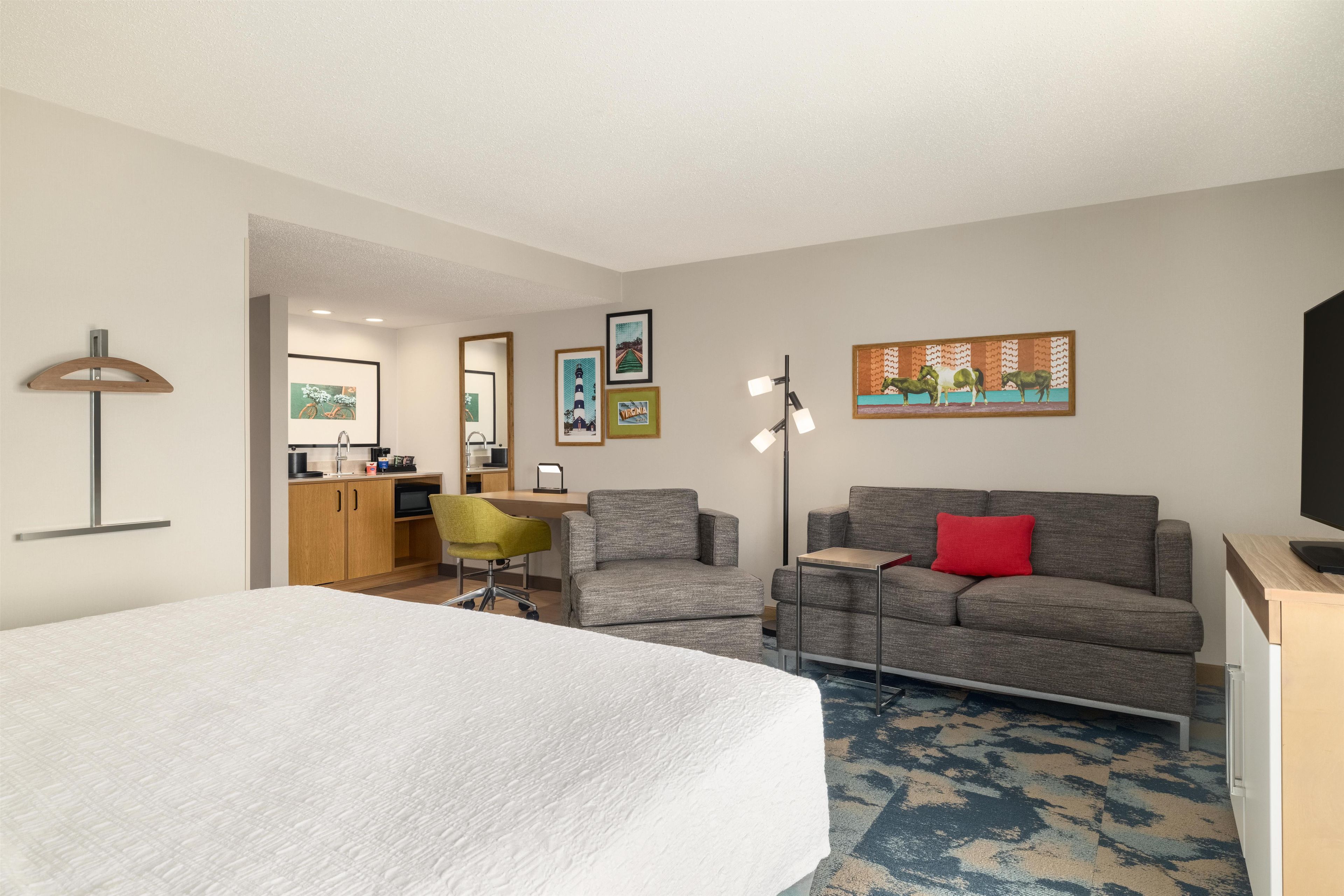 A hotel room with a bed, a sofa, a desk, and a chair. There are framed pictures on the wall and a lamp.