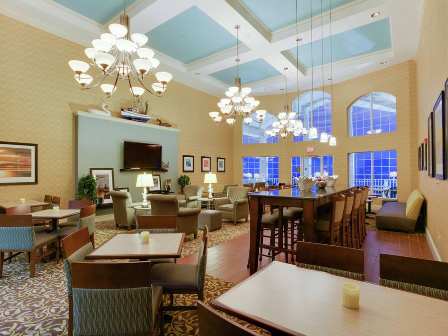 A hotel lobby with multiple seating areas, including tables and chairs, a television, and large windows allowing natural light.