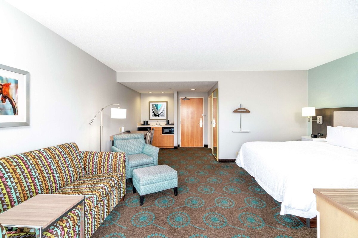 A hotel room with a bed, a sofa, a chair, a small table, a desk, a lamp, a microwave, a cabinet, and a door leading to a bathroom.
