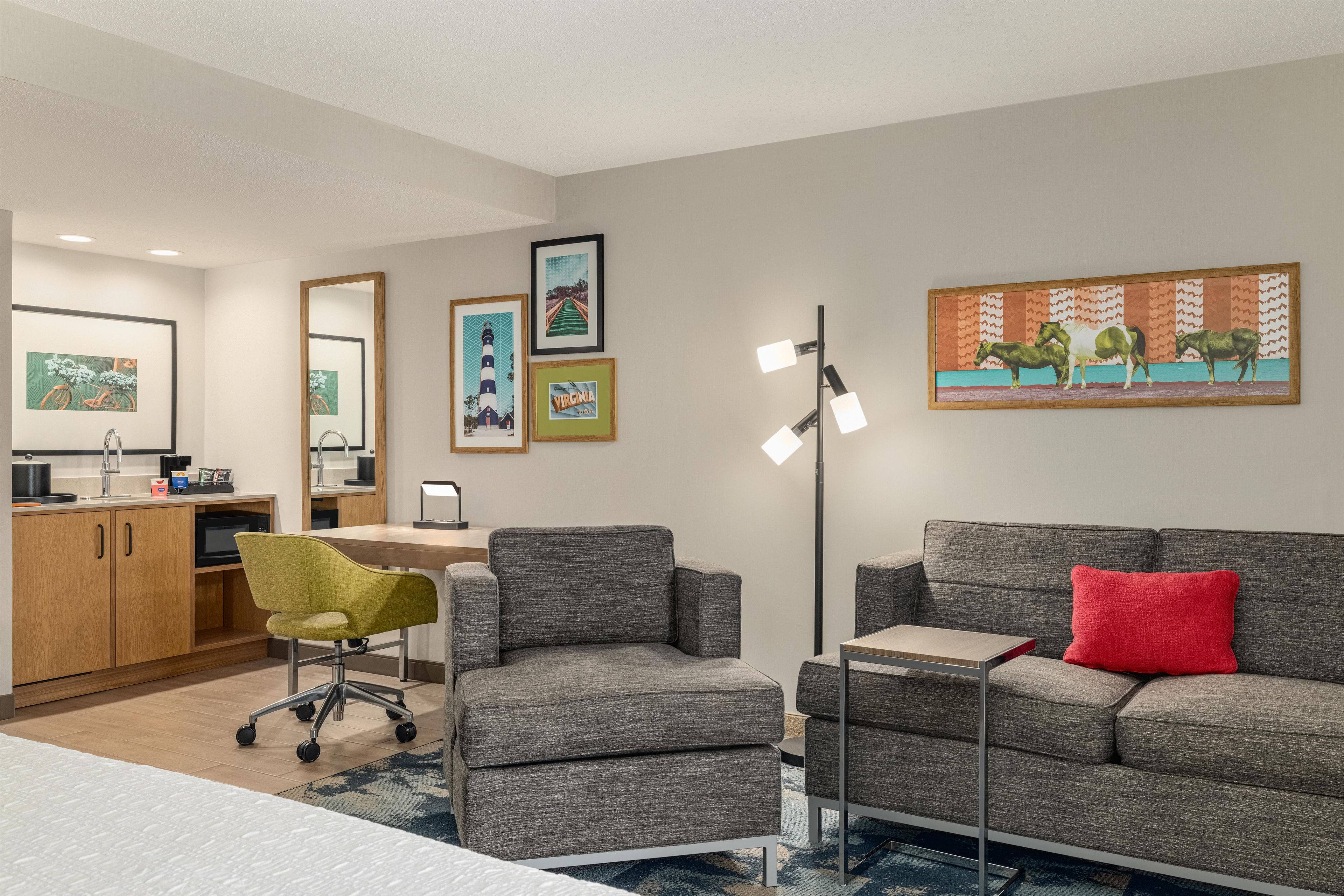 A modern living room with a grey sofa, a red pillow, a green office chair, and a desk with a computer. There are framed pictures on the wall and a floor lamp.