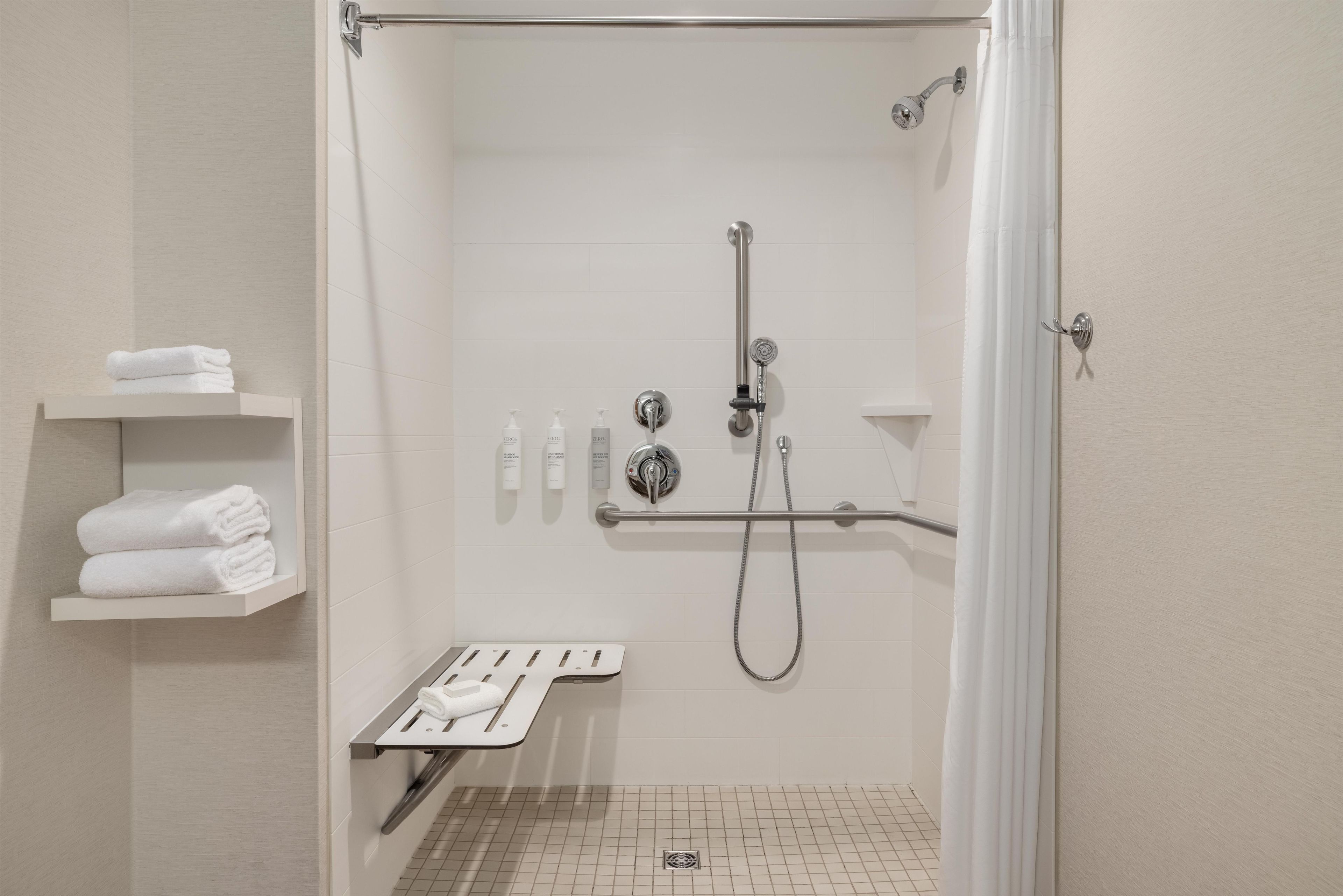 Un espace douche moderne doté d’un pommeau de douche à main, d’un flexible de douche et d’un robinet mural. Trois serviettes blanches sont pliées et empilées sur une étagère située à gauche.