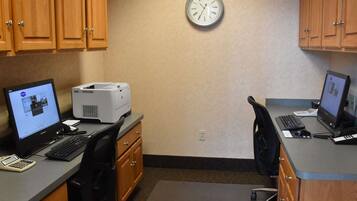 A small office with two desks, each with a computer, keyboard, and mouse. There is a printer on one desk, and a clock on the wall.