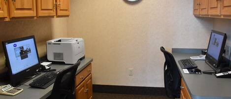 A small office with two desks, each with a computer, keyboard, and mouse. There is a printer on one desk, and a clock on the wall.