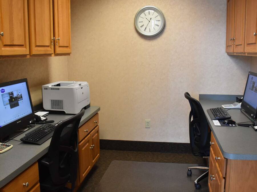 A small office with two desks, each with a computer, keyboard, and mouse. There is a printer on one desk, and a clock on the wall.