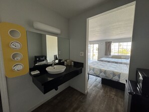 A hotel room with a black countertop, a white sink, a mirror, and a bed with patterned bedding. - Motel 6 Delano, CA (Delano)