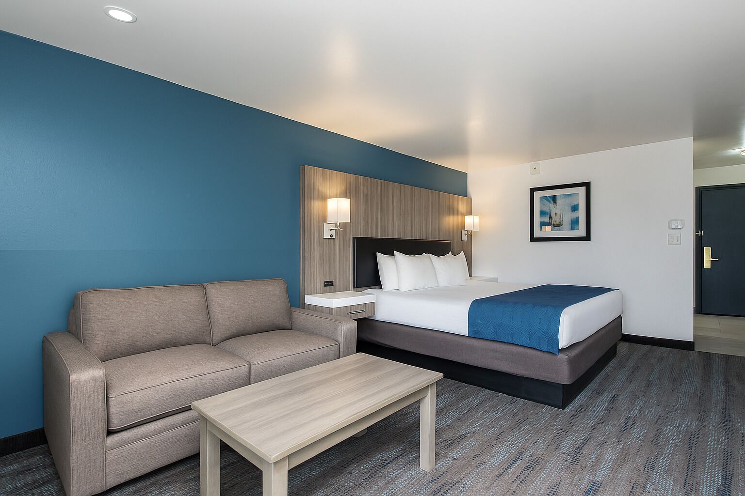 Una habitación de hotel con cama, sofá, una mesita, televisor y un cuadro en la pared.