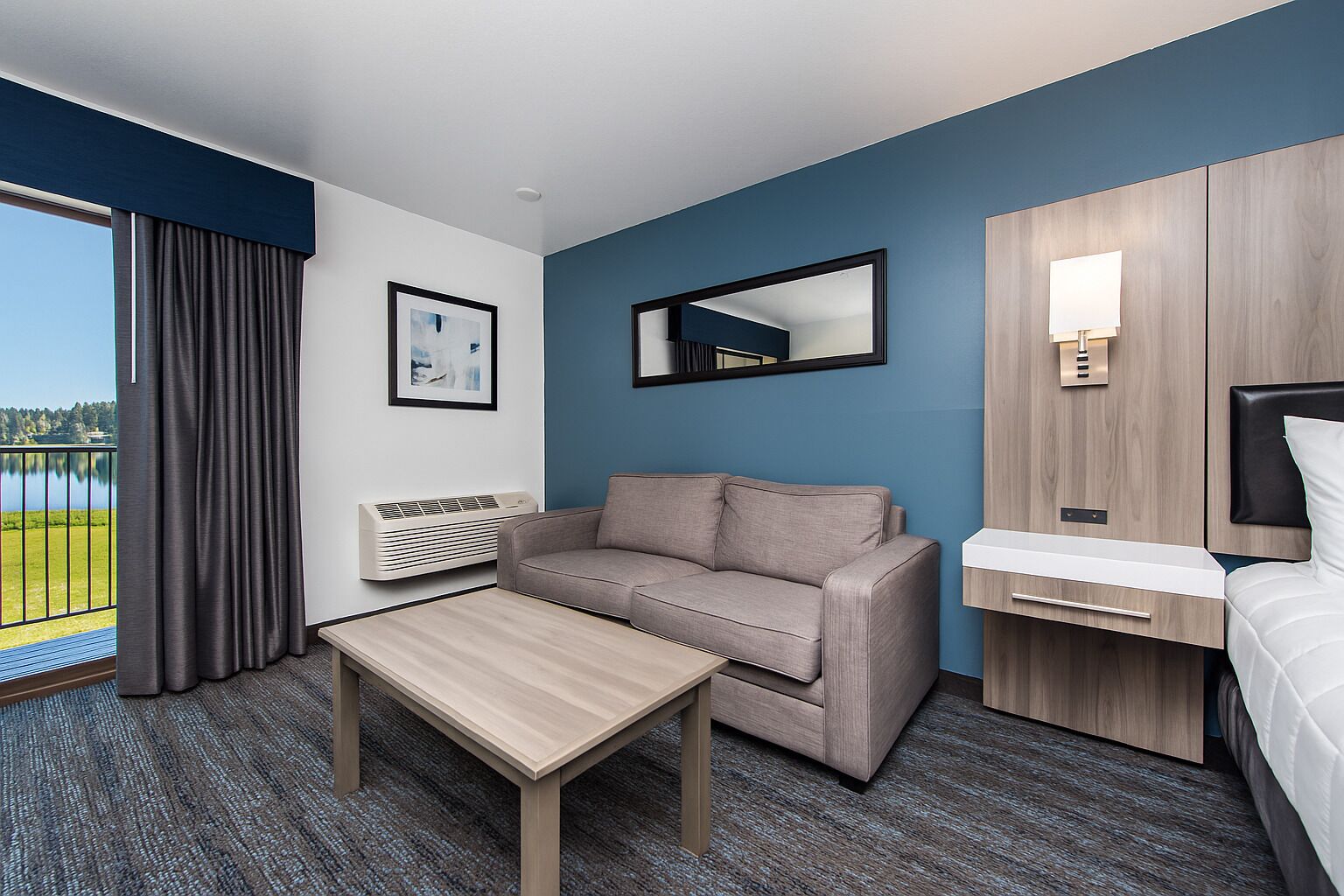Habitación de hotel con sofá, una mesita, cama, espejo y ventana con vista a un lago.