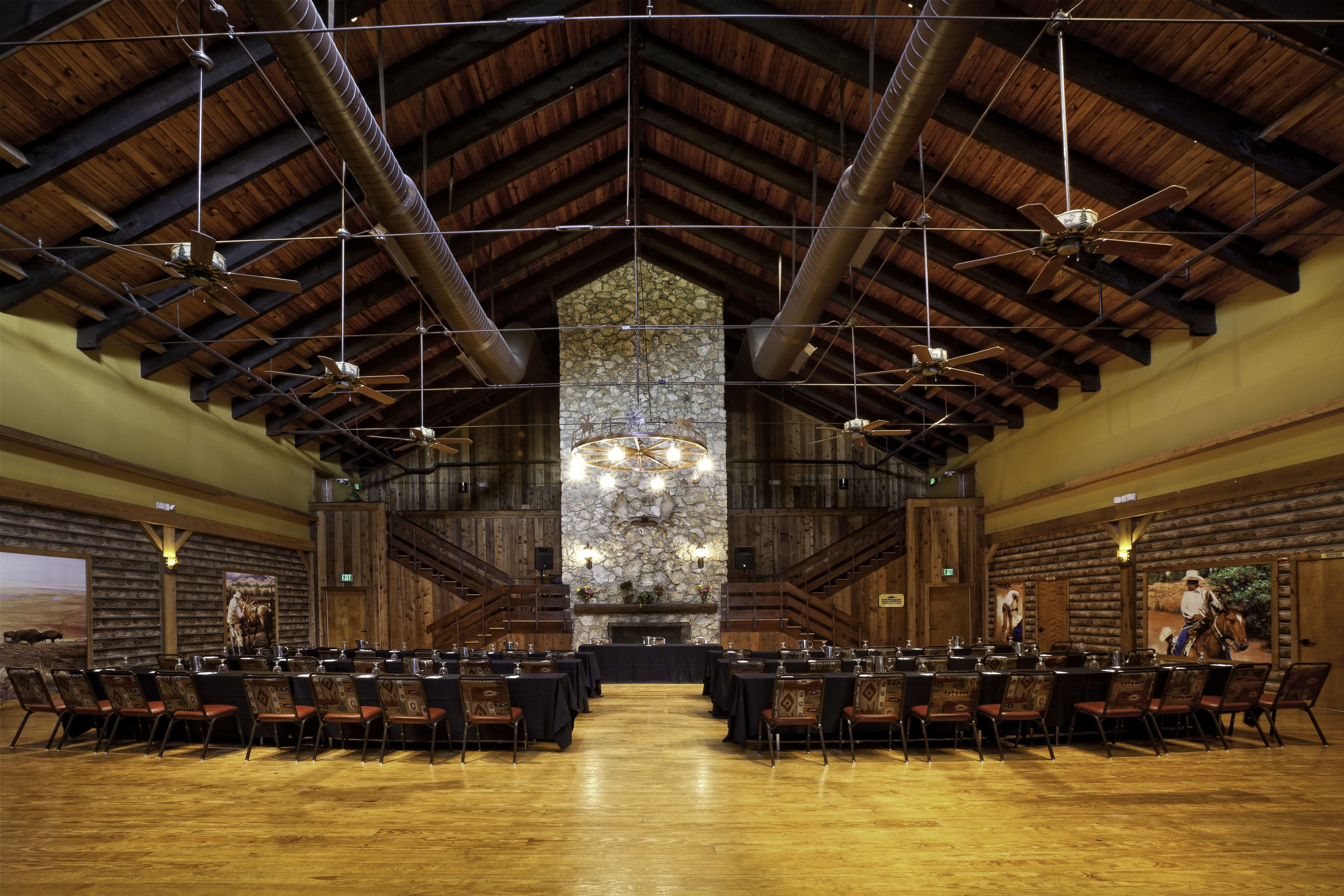 A large hall with high ceilings, wooden beams, and a central stone fireplace. Tables and chairs are arranged for dining.