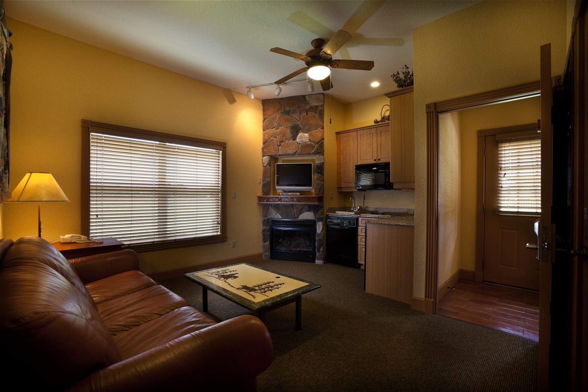 A living room with a brown leather couch, a coffee table, a fireplace, and a kitchen area with wooden cabinets.