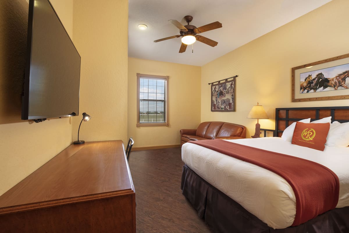 Une chambre d’hôtel comprenant un lit, un bureau, une télévision, un ventilateur de plafond, une fenêtre avec des stores, un tableau au mur et une lampe.