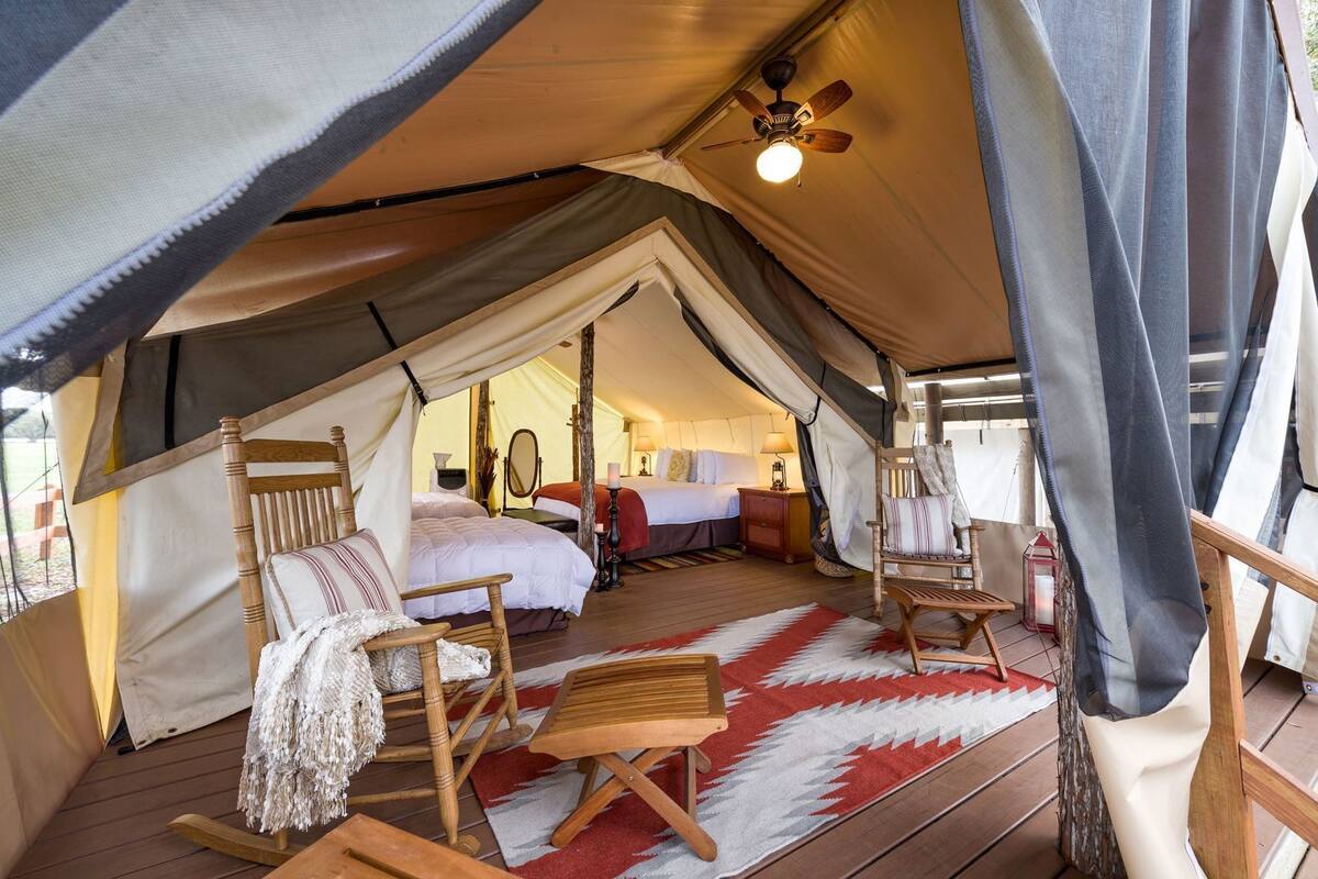 Un hébergement sous tente comprenant un lit, deux chaises, un fauteuil à bascule, un ventilateur de plafond et un tapis rouge et blanc.