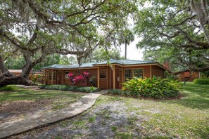 A rustic cabin with a green roof, surrounded by lush greenery and large trees with Spanish moss.