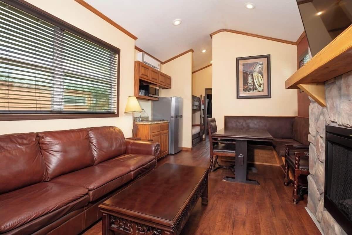 Un salon avec un canapé en cuir marron, une table basse en bois, une table à manger avec des chaises, une cheminée en pierre et un coin cuisine avec des placards et des appareils électroménagers.