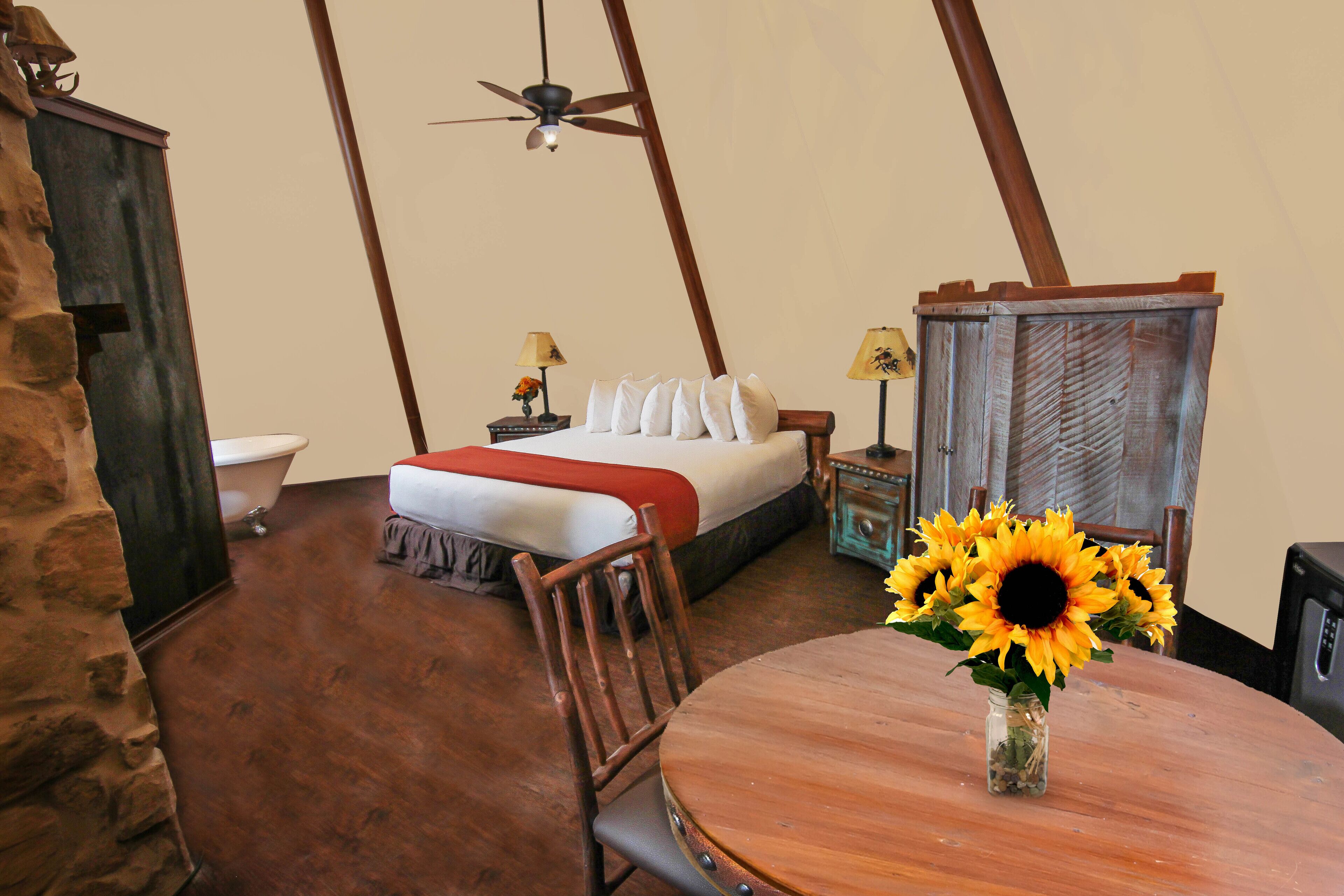 A room with a bed, a wooden table with a vase of sunflowers, a ceiling fan, and a stone wall.