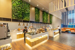 Un café moderne avec un comptoir central, un espace salon doté de coussins orange et des murs végétalisés.