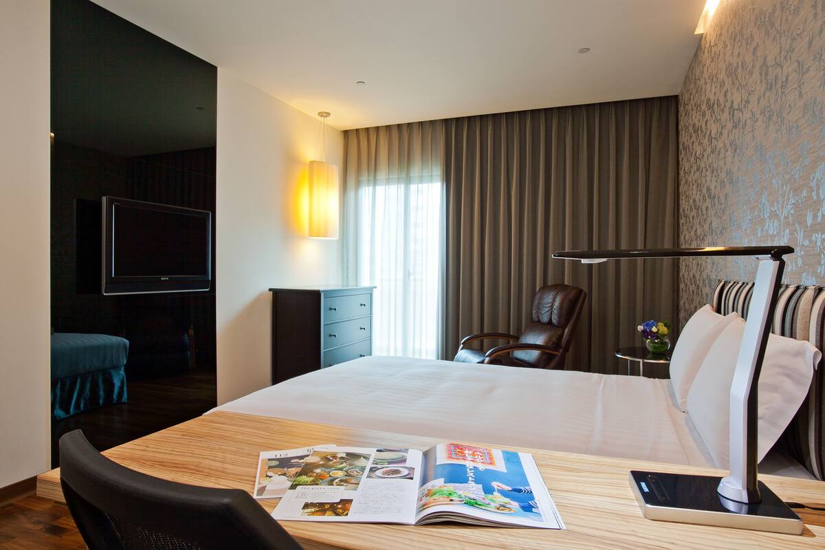 Habitación de hotel con una cama grande, un escritorio con una revista, una silla, un televisor y una lámpara.