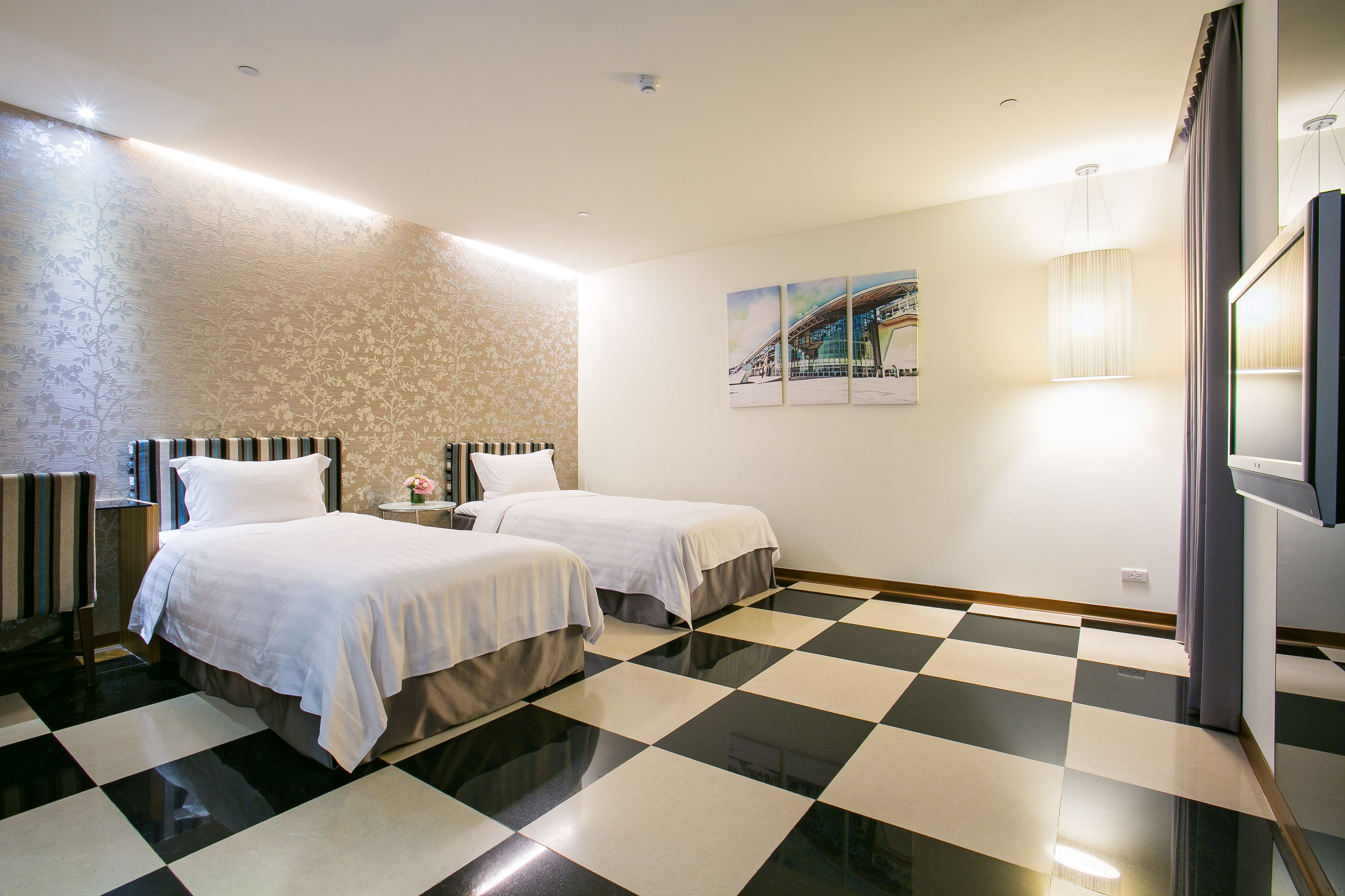 A hotel room with two beds, a checkered floor, a patterned wall, and a painting.