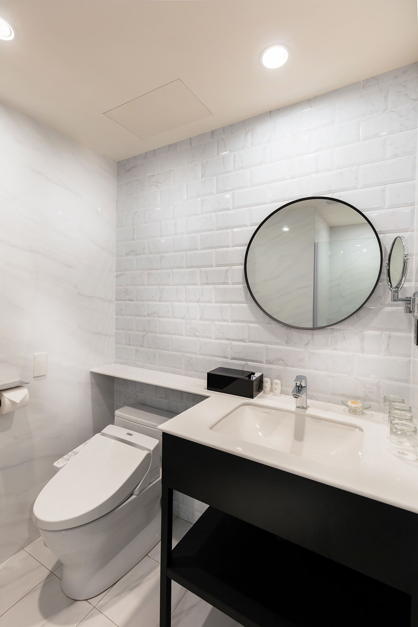 A modern bathroom with a white toilet, a black vanity, a round mirror, and a marble-tiled wall.