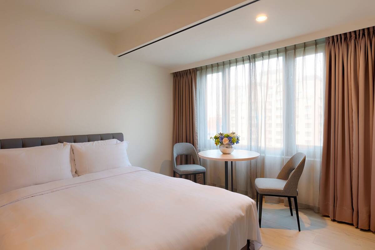 Una habitación de hotel con una cama grande, una mesa redonda con dos sillas y una ventana con cortinas.