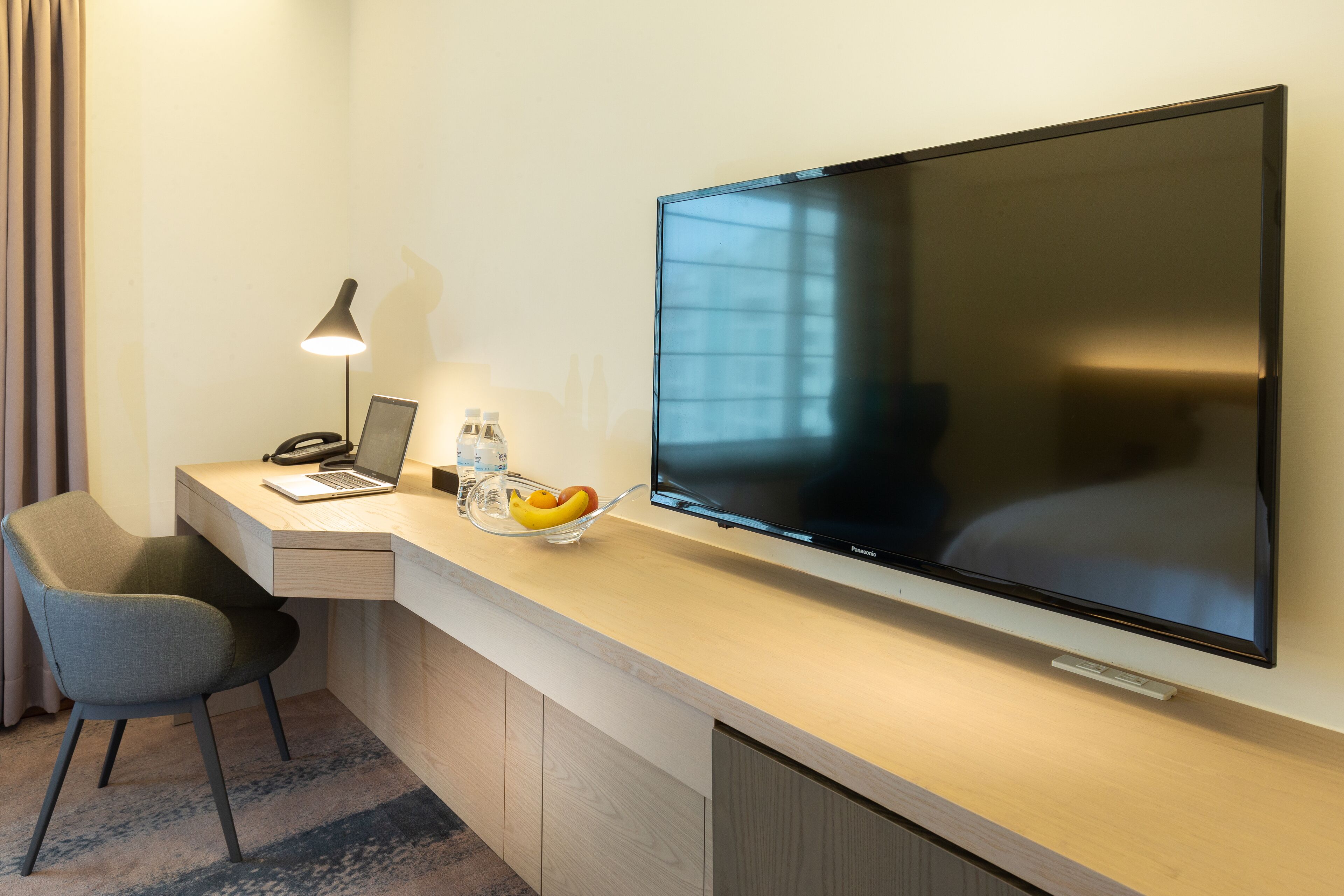 A hotel room with a desk, a laptop, a flat-screen TV, a chair, a bowl of fruit, and a lamp.