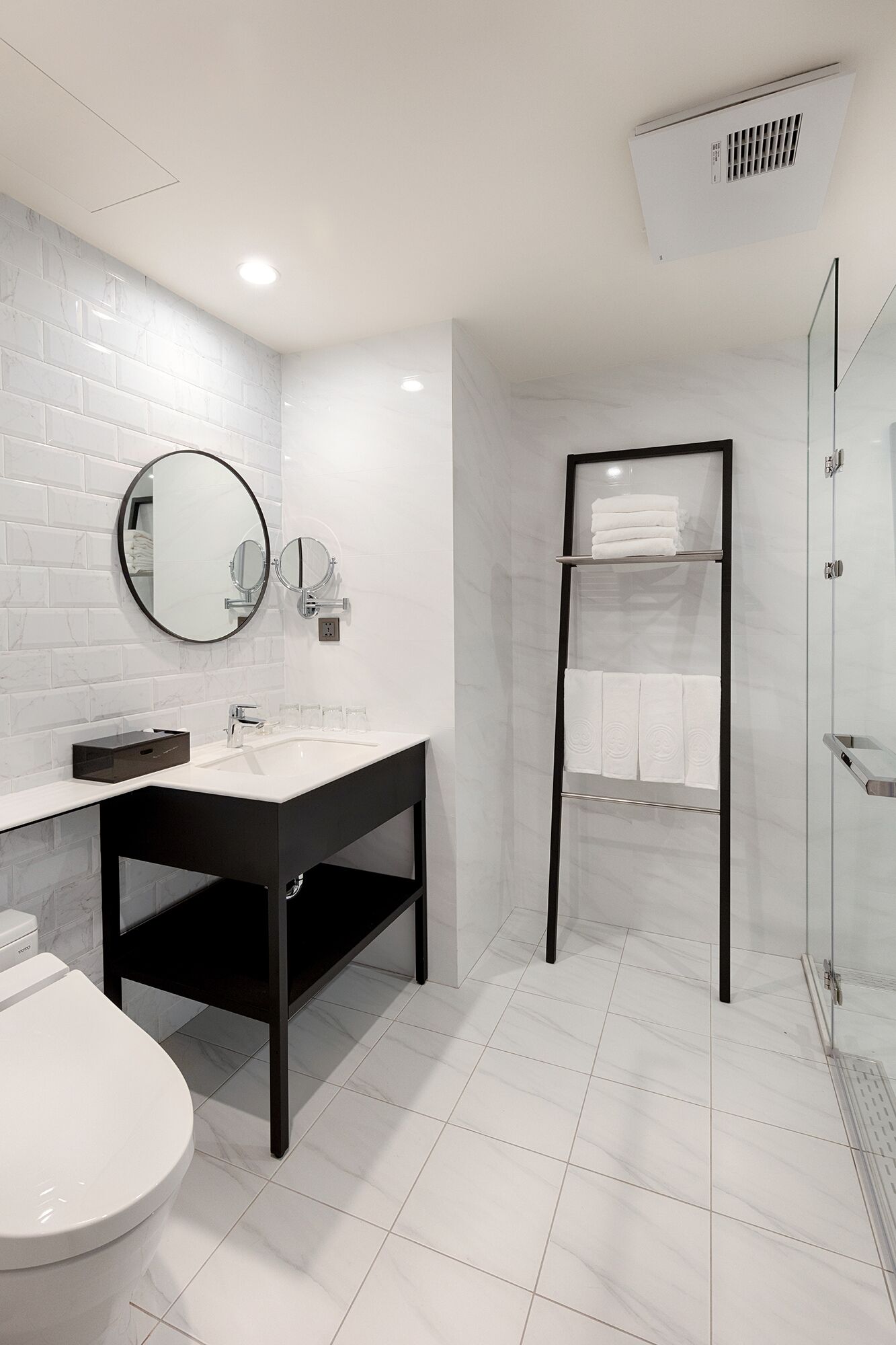 A modern bathroom with a white tiled floor, a black vanity with a sink, a toilet, a glass shower enclosure, and a towel rack with folded towels.