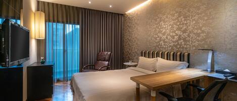 Habitación de hotel con una cama grande, un escritorio, una silla, un televisor y un ventanal con cortinas.