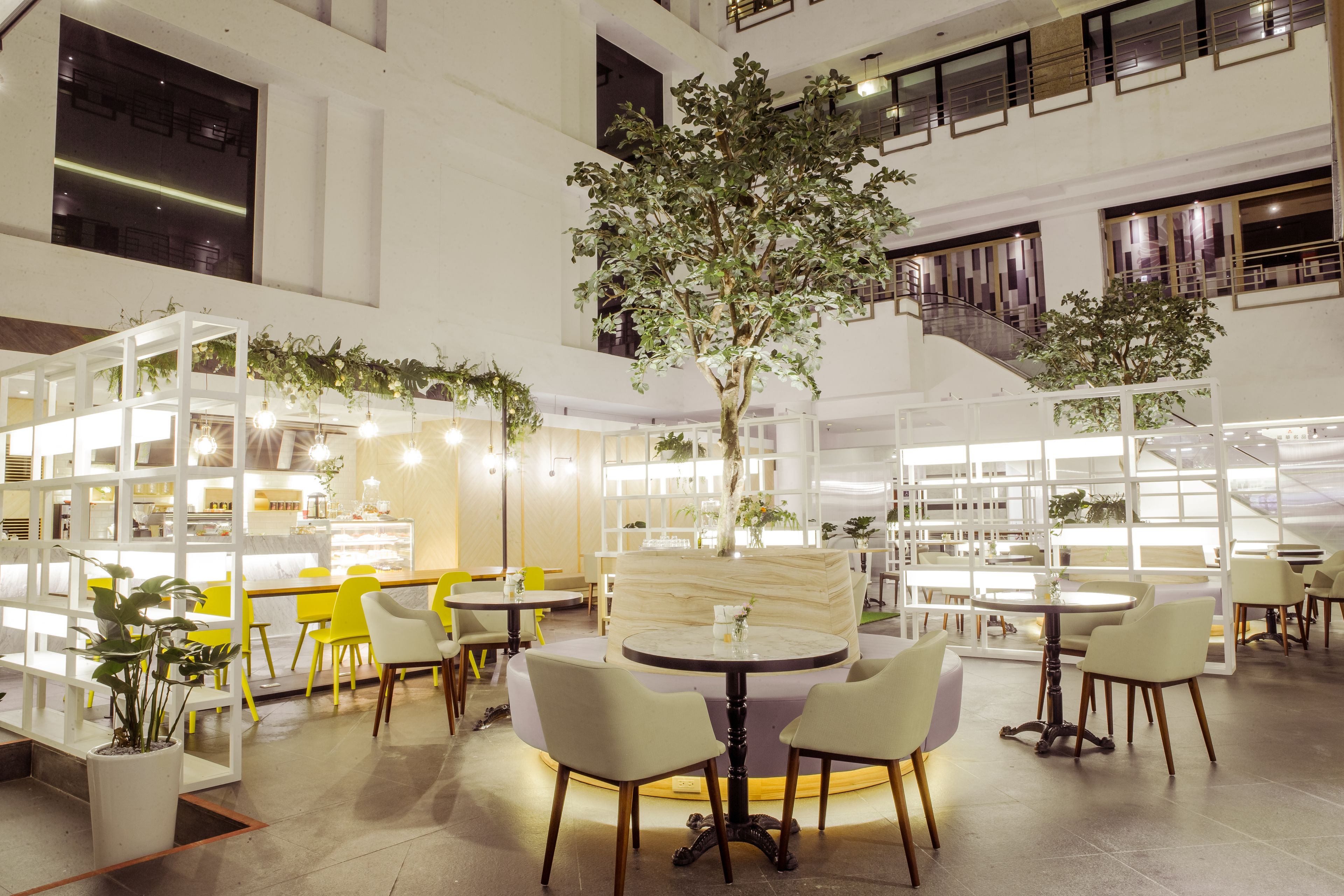 Un espacio interior amplio con varias mesas y sillas, un árbol central y un diseño arquitectónico moderno con paredes blancas y detalles de madera.