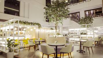Un espacio interior amplio con varias mesas y sillas, un árbol central y un diseño arquitectónico moderno con paredes blancas y detalles de madera.