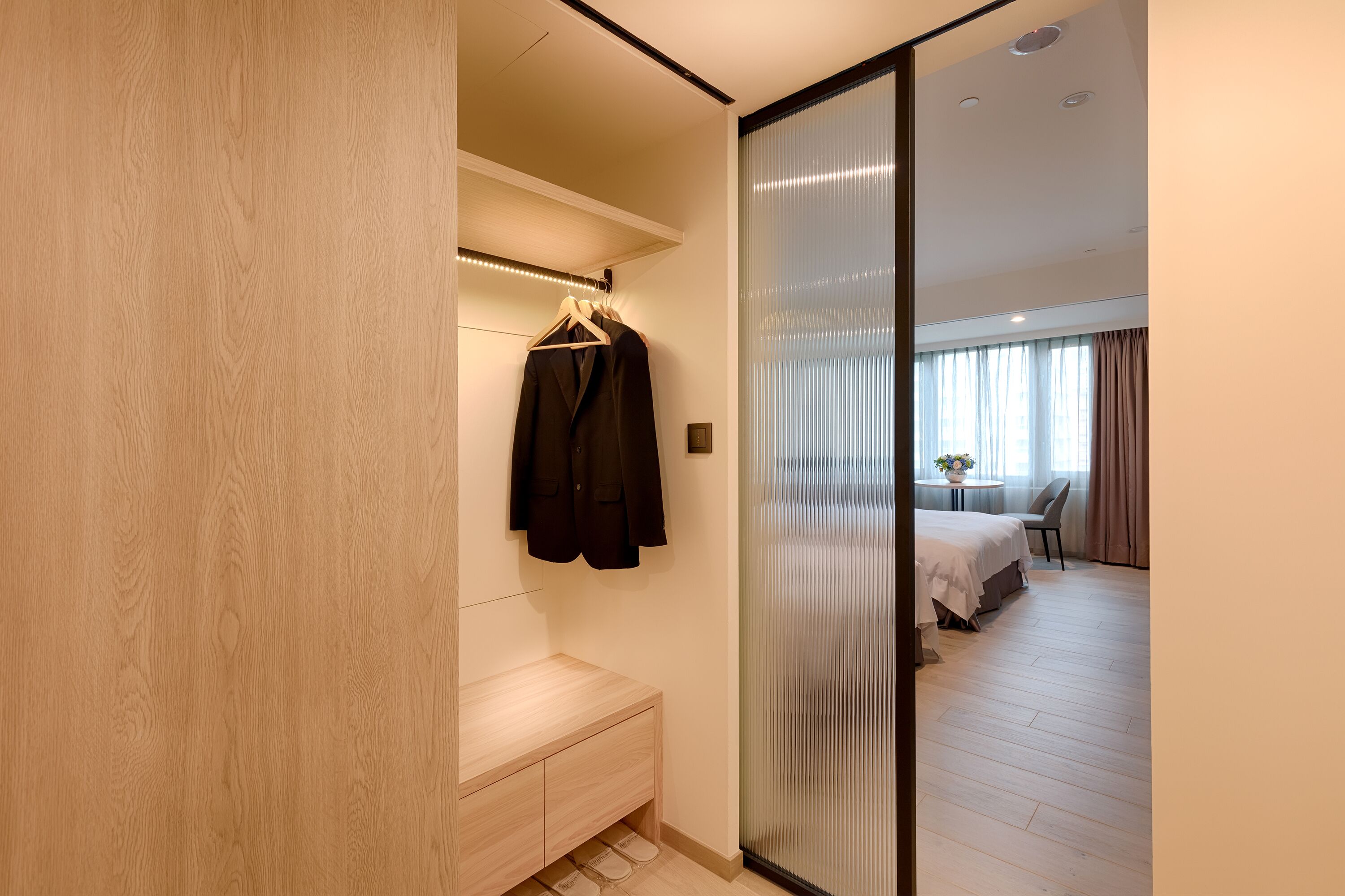 Una habitación de hotel moderna con un banco de madera, un perchero con una chaqueta negra y un ventanal corredero que da a un dormitorio con una cama y una mesita.