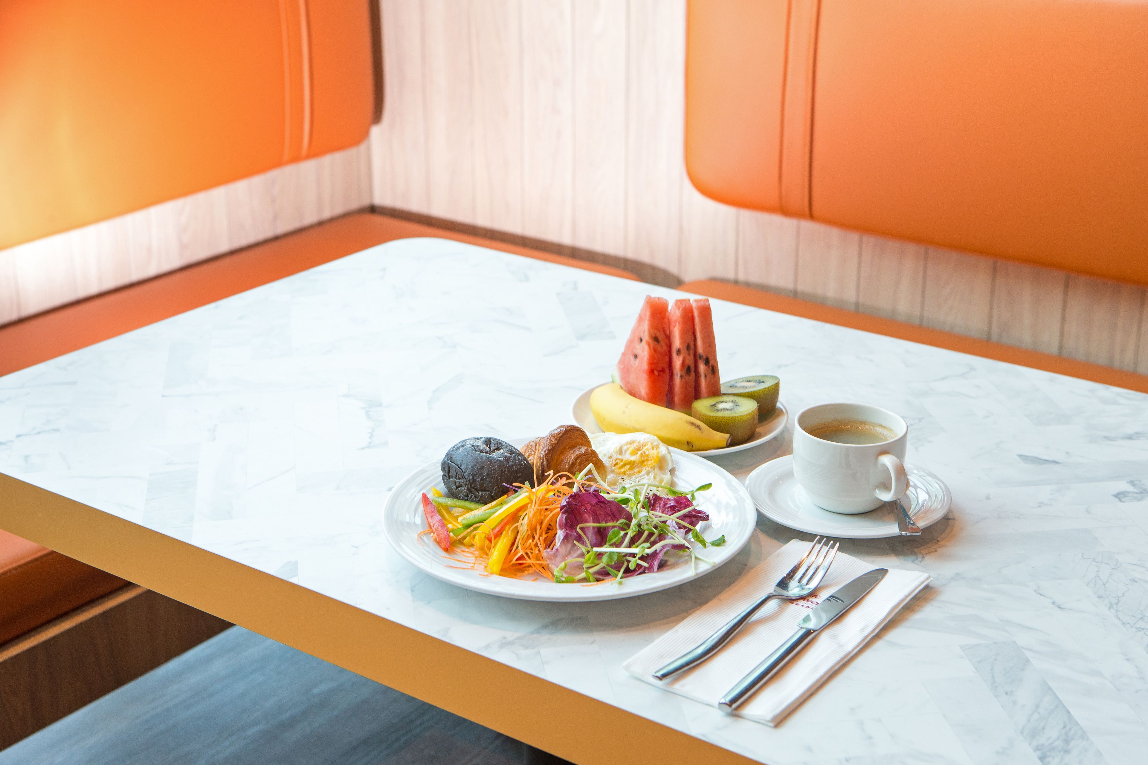 A dining table with a plate of food, a banana, a kiwi, a slice of watermelon, and a cup of coffee.