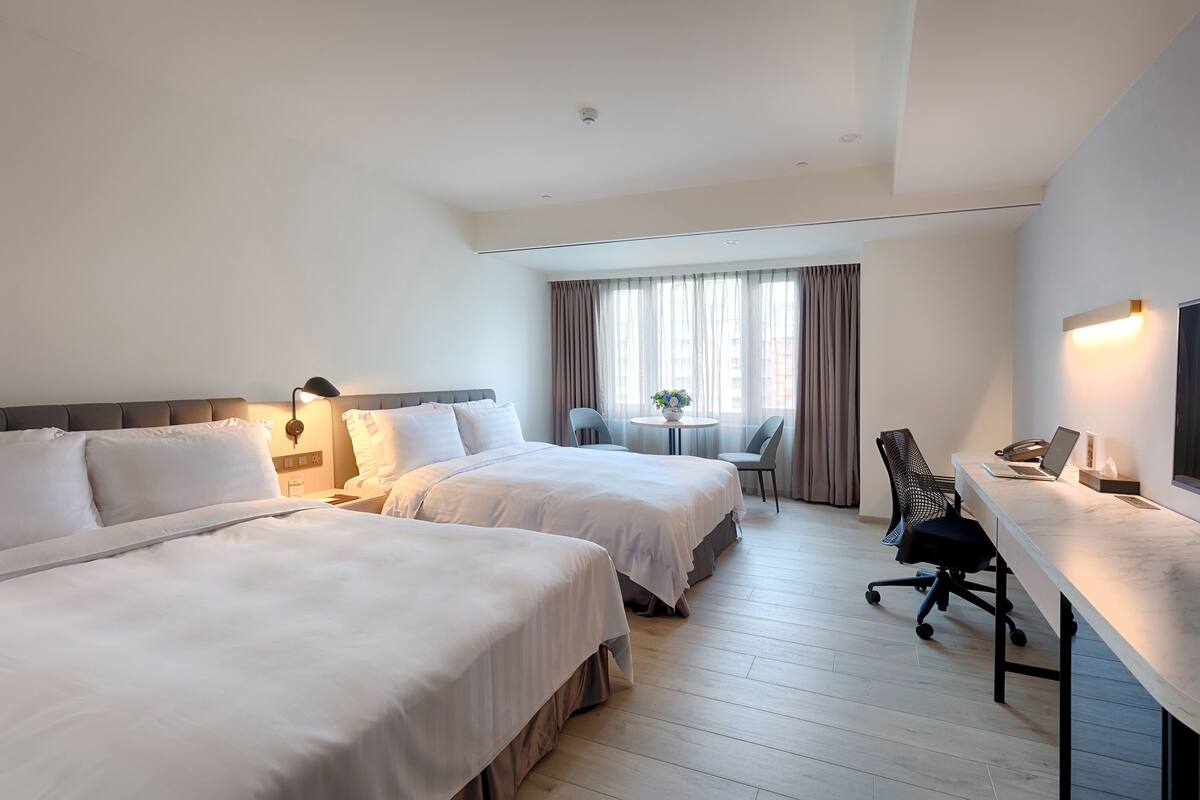 Habitación de hotel con dos camas, un escritorio con una computadora portátil, una silla, una mesita de noche con un florero y una ventana con cortinas.