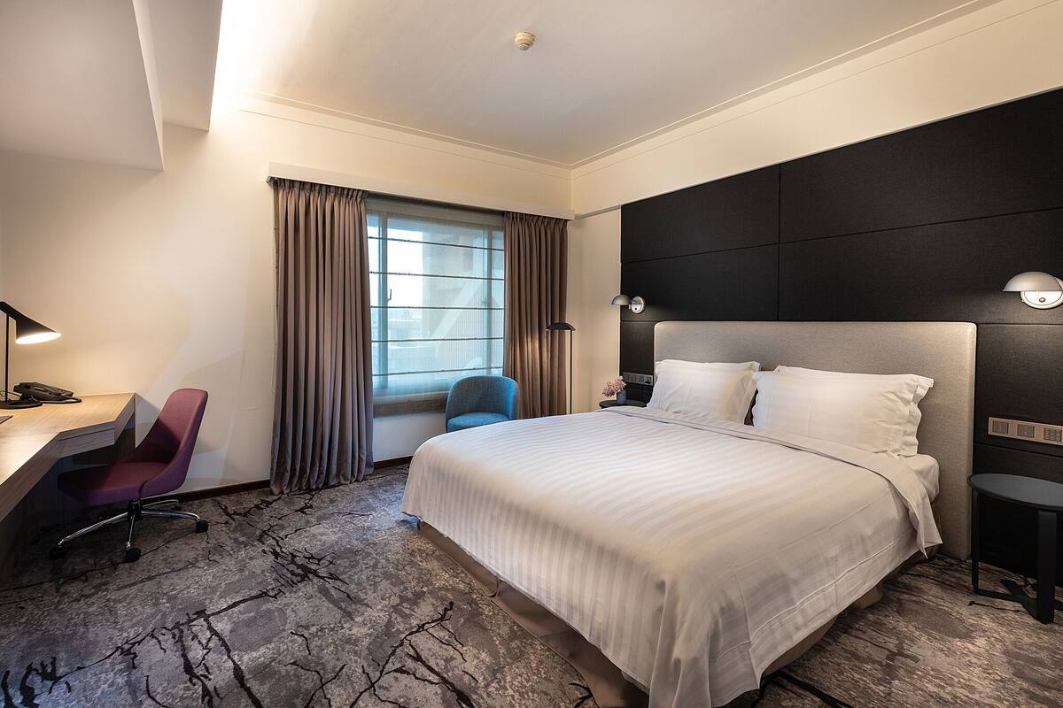 Habitación de hotel con una cama grande, un escritorio con silla, una mesita pequeña y una ventana con cortinas.