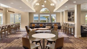 A modern hotel lobby with a high ceiling, large windows, a seating area with sofas and chairs, and a bar with stools. - Residence Inn by Marriott Sacramento Folsom (Folsom)