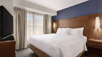 A hotel room with a large bed, a flat-screen TV on a wooden stand, a nightstand with a lamp, and a view of the city through the window.