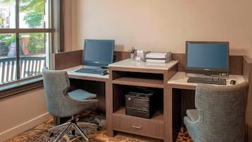 A workspace with two desks, each with a computer and keyboard, and a printer. There are two office chairs and a small shelf with office supplies.