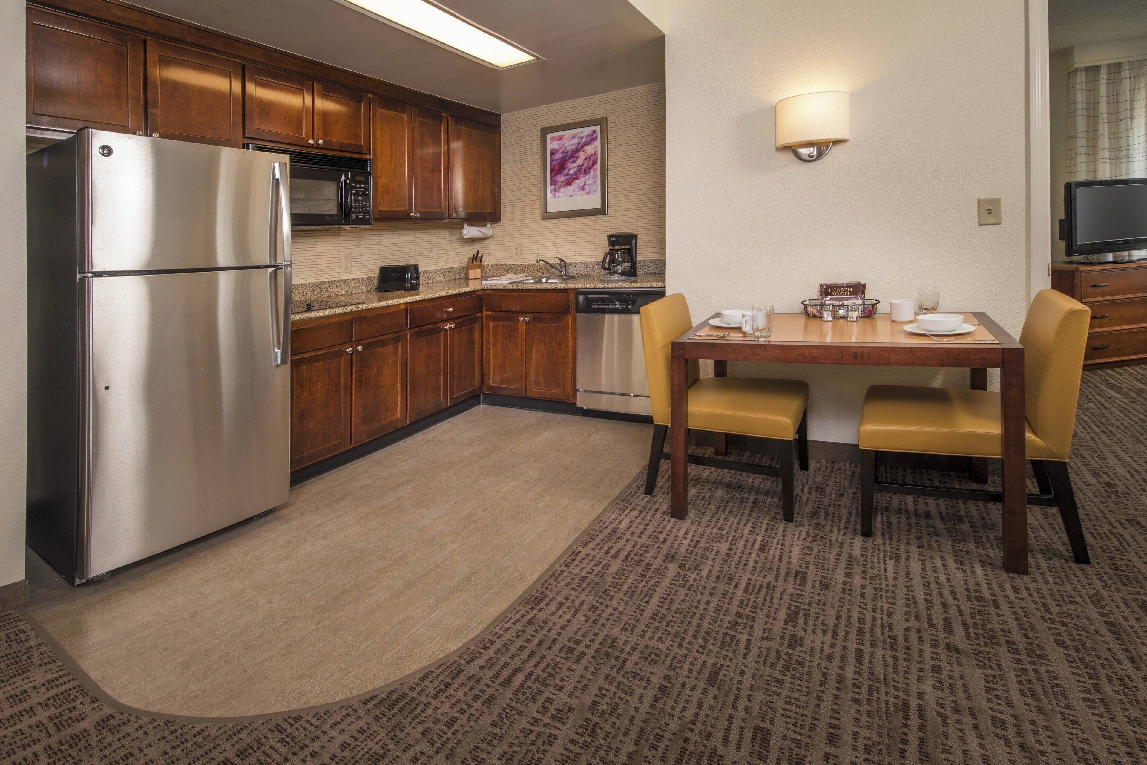 Cocina compacta con refrigerador, microondas y cafetera. Cuenta con una mesa de comedor con sillas y un televisor pequeño.