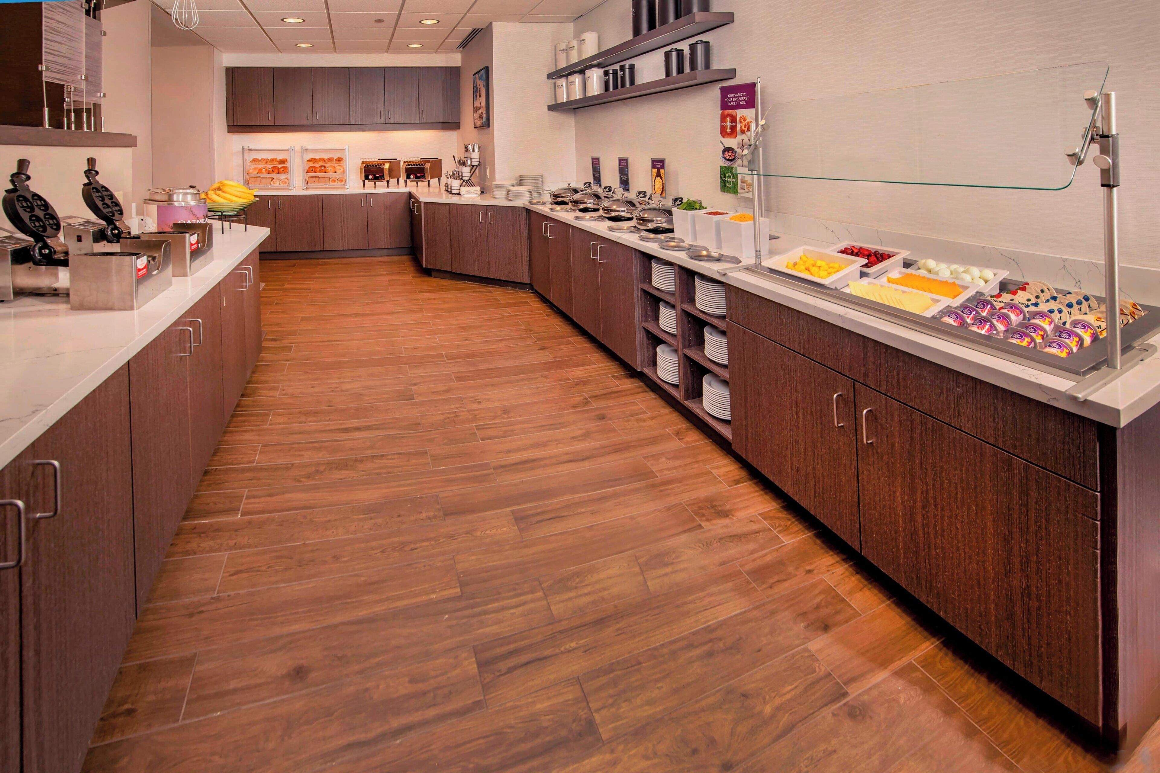 A modern hotel kitchen with wooden flooring, stainless steel appliances, and a buffet area with various food items.