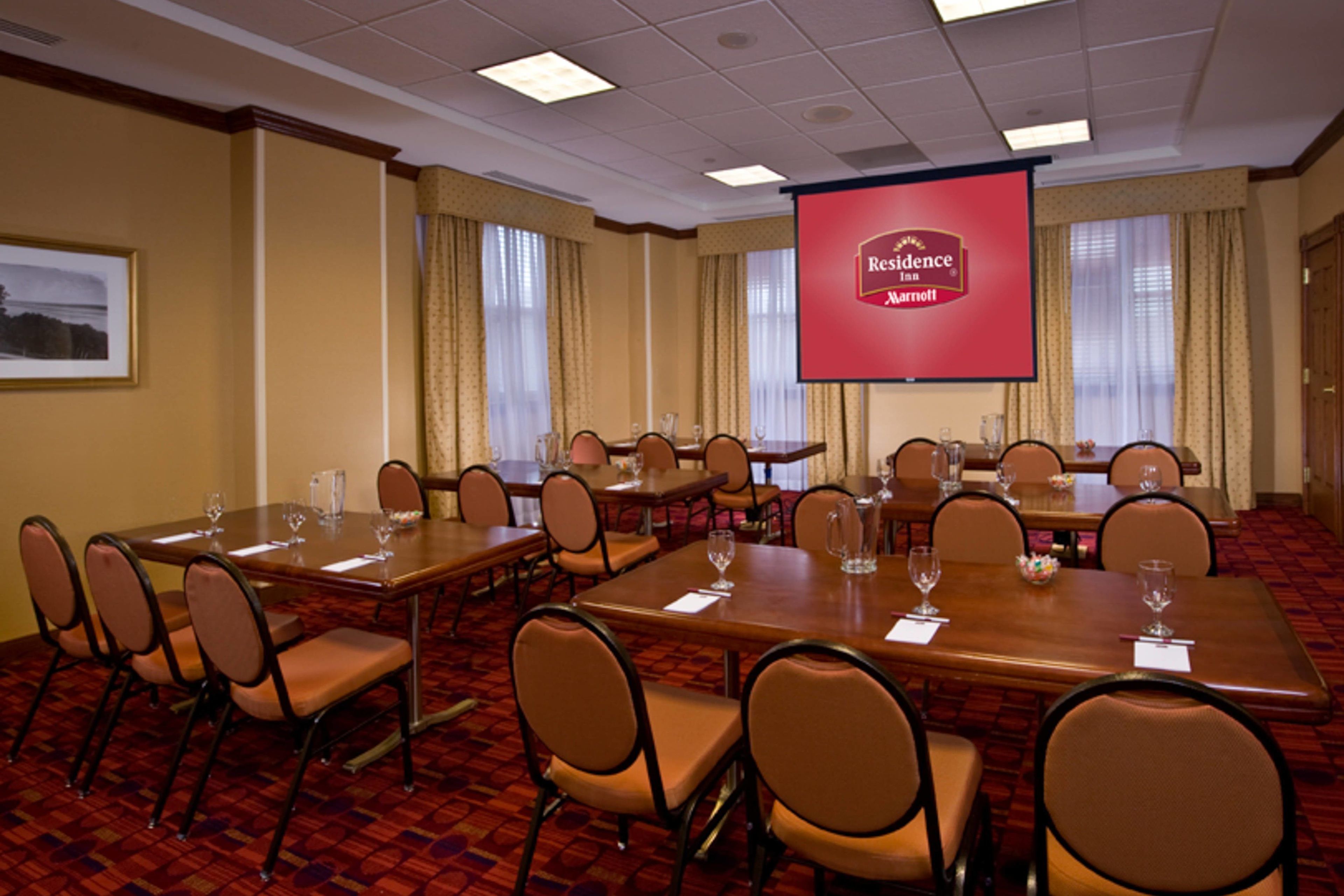 Un salón de conferencias con varias mesas y sillas, una pantalla grande que muestra el logotipo de Residence Inn Marriott, y un cuadro en la pared.