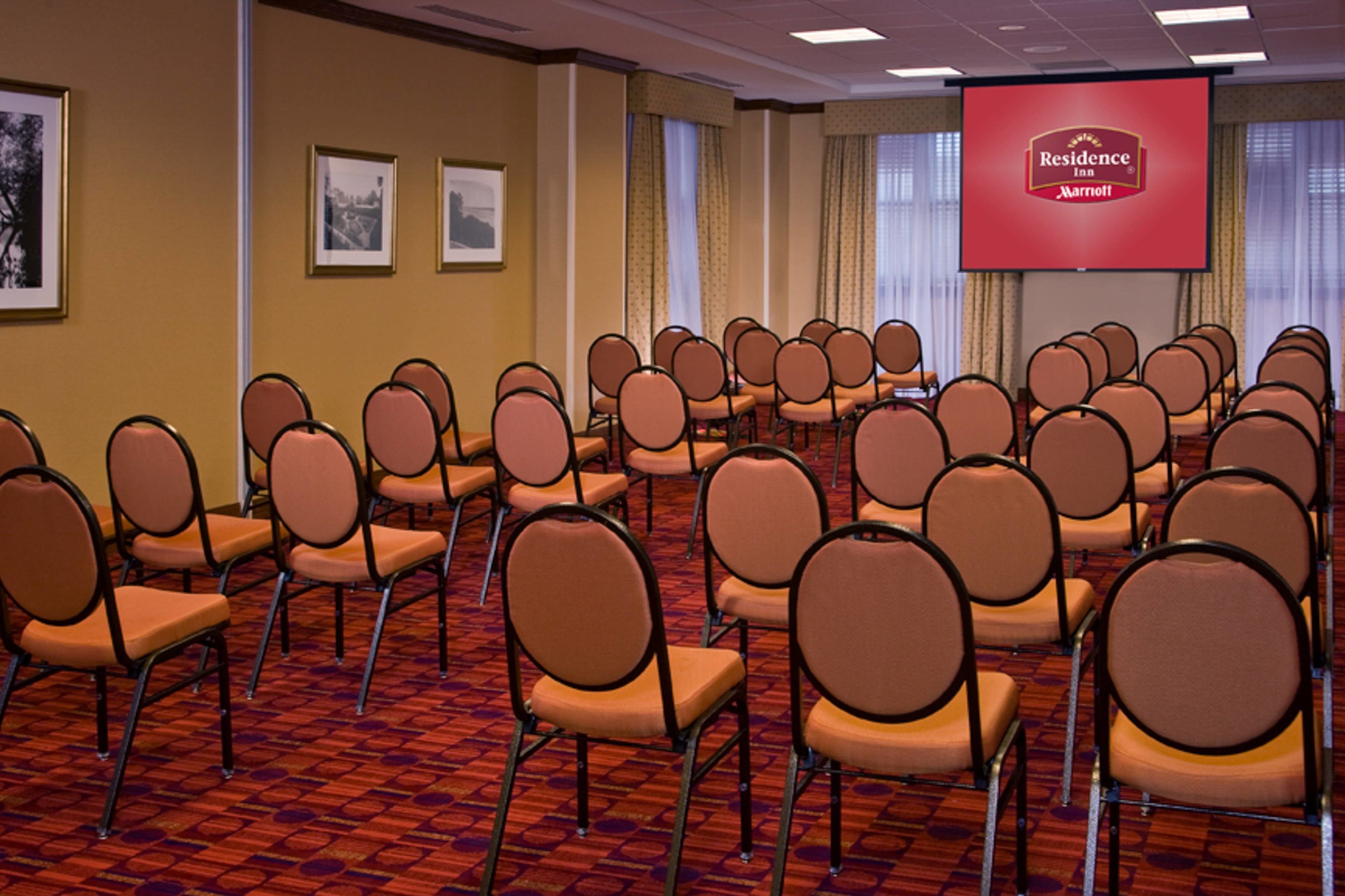 Uma sala de conferência com fileiras de cadeiras, uma tela grande exibindo o logotipo do Residence Inn Marriott, quadros nas paredes e um tapete estampado.