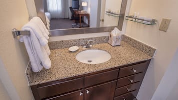 Une salle de bain avec un lavabo doté d’un plan de travail en granit, un miroir, un porte-serviettes et une étagère en verre où sont rangés les articles de toilette.
