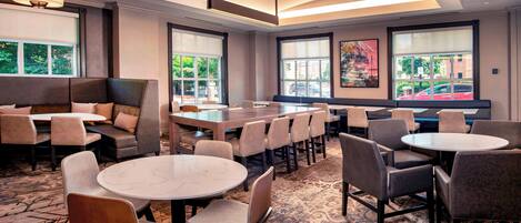 Un hall d’hôtel avec des tables rondes, des canapés et des fauteuils disposés pour permettre aux clients de prendre un repas ou de se détendre. De grandes fenêtres offrent une vue sur l’extérieur.